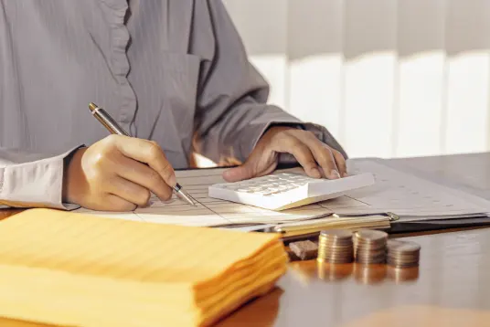 Young student at desk using calculator and writing budget calculations on paper with coins and bills visible