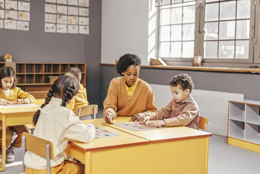 Group of engaged students around table playing educational card game with teacher supervising in classroom setting